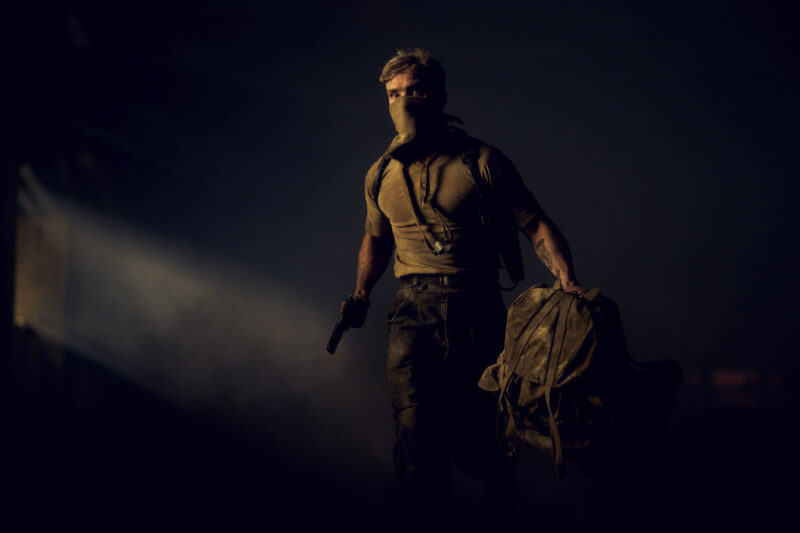 "A masked man with a gun and a duffel bag walks through a dark, smoky environment with a beam of light shining in the background, suggesting a tense or action-packed setting."