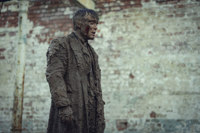 "A man covered in thick mud stands outdoors in front of a weathered brick wall, wearing tattered, muddy clothing. The setting appears gritty and industrial, suggesting harsh conditions or an intense situation."