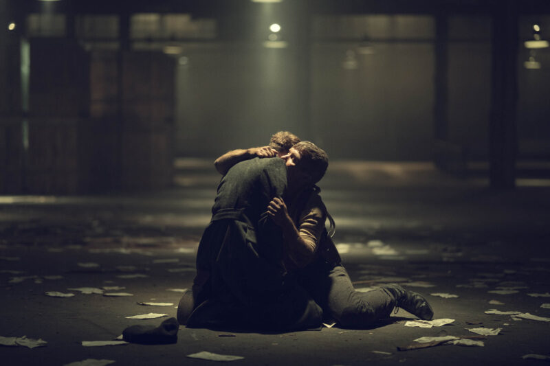 "Two people kneel on the floor of a dimly lit warehouse, embracing each other, surrounded by scattered papers and crates in a dramatic, emotional setting."