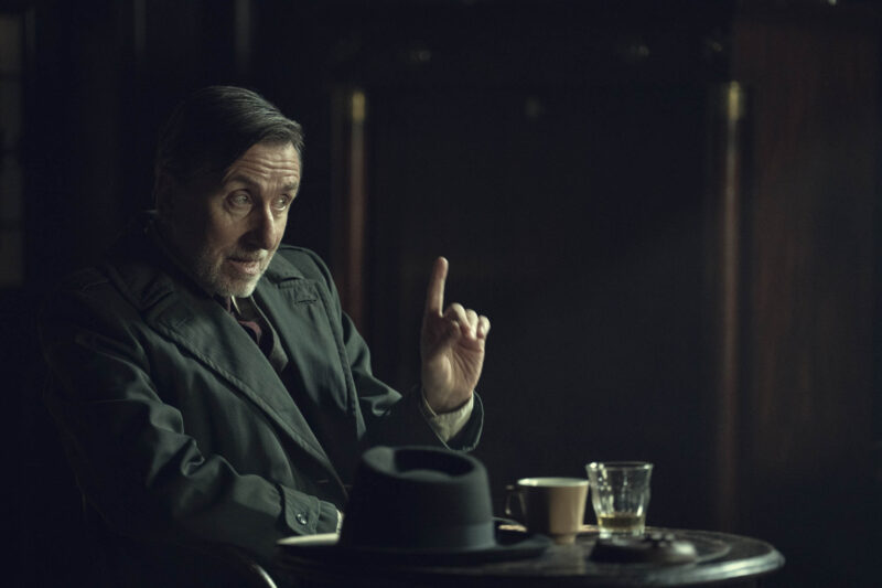 "Older man in a dark suit sits at a small round table in a dimly lit room, gesturing with his finger raised. Hat, coffee cup, and glass are on the table, suggesting a serious conversation or meeting in a moody cafe or bar."