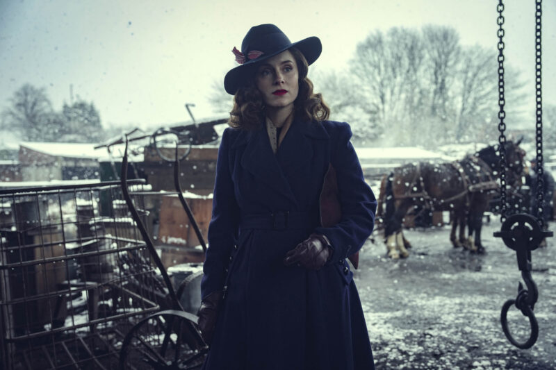 "Woman in vintage coat and hat standing outdoors in snowy, wintry setting, with old carts and horses in the background, evoking a historical or period atmosphere."