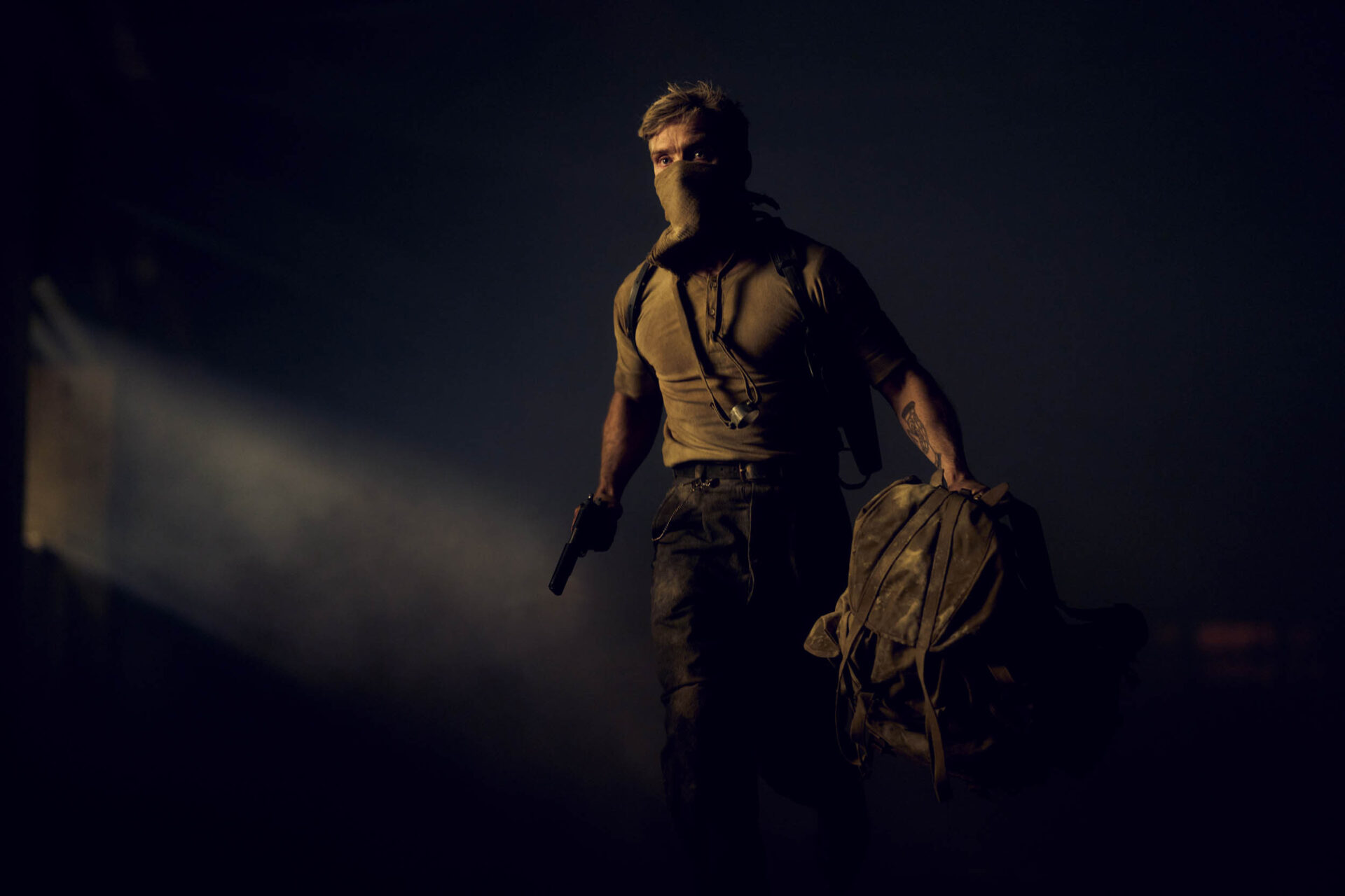 "A masked man with a gun and a duffel bag walks through a dark, smoky environment with a beam of light shining in the background, suggesting a tense or action-packed setting."