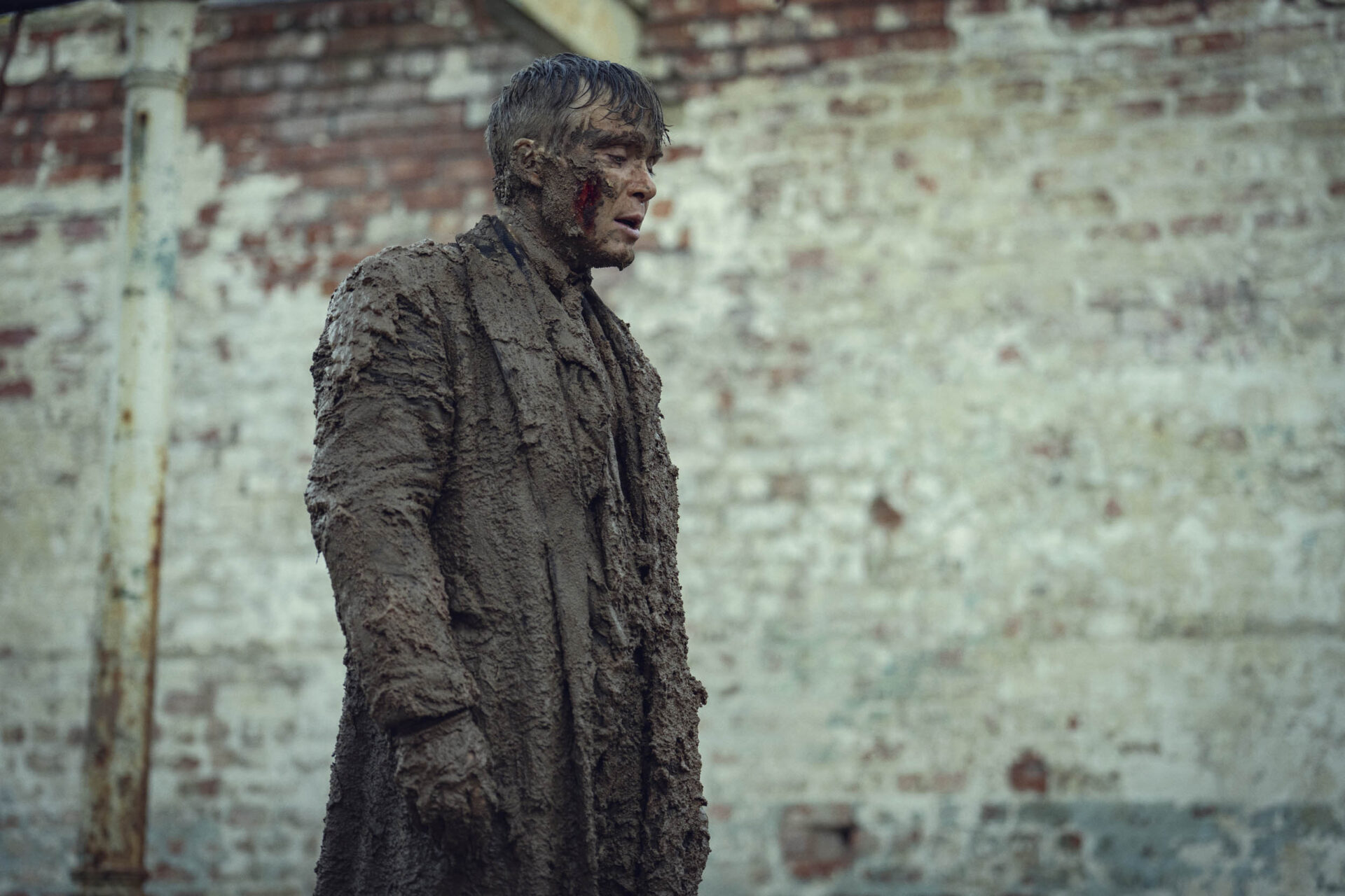 "A man covered in thick mud stands outdoors in front of a weathered brick wall, wearing tattered, muddy clothing. The setting appears gritty and industrial, suggesting harsh conditions or an intense situation."