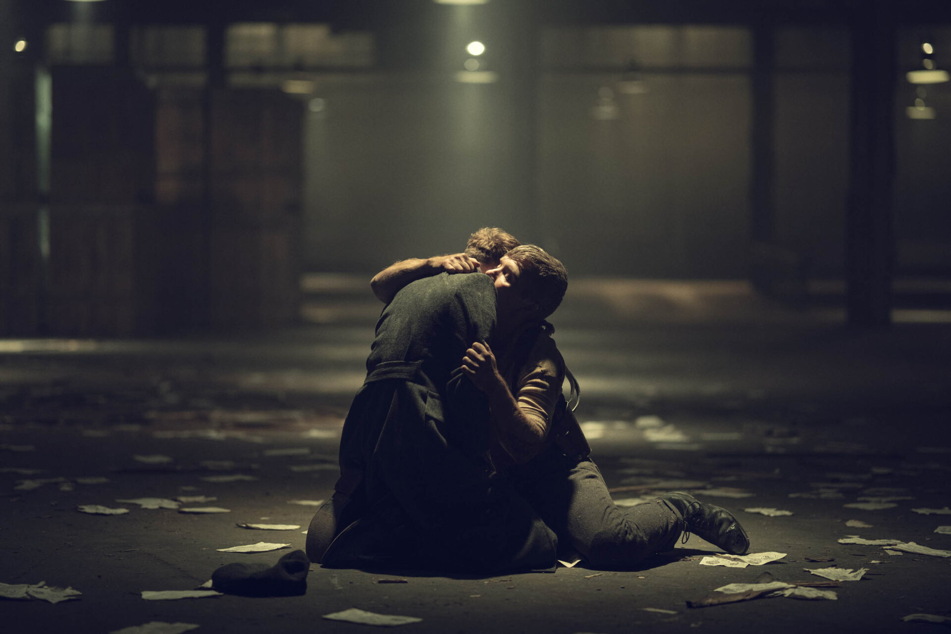 "Two people kneel on the floor of a dimly lit warehouse, embracing each other, surrounded by scattered papers and crates in a dramatic, emotional setting."