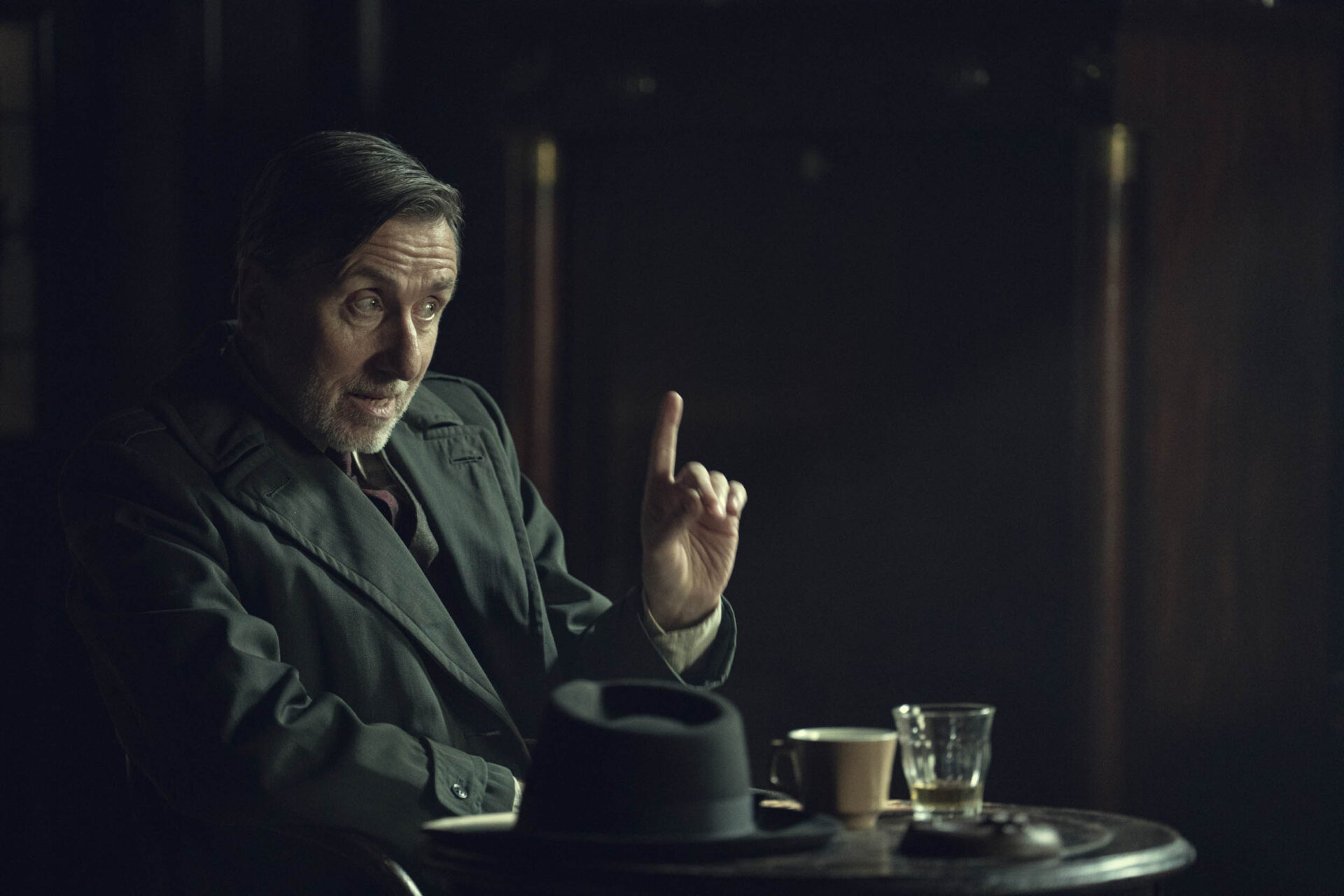 "Older man in a dark suit sits at a small round table in a dimly lit room, gesturing with his finger raised. Hat, coffee cup, and glass are on the table, suggesting a serious conversation or meeting in a moody cafe or bar."