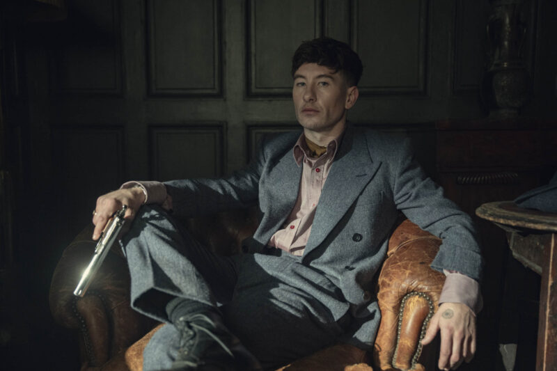 "Man in a gray suit sitting in a vintage leather armchair, holding a gun, in a dimly lit room with dark paneled walls, creating a tense and dramatic atmosphere."