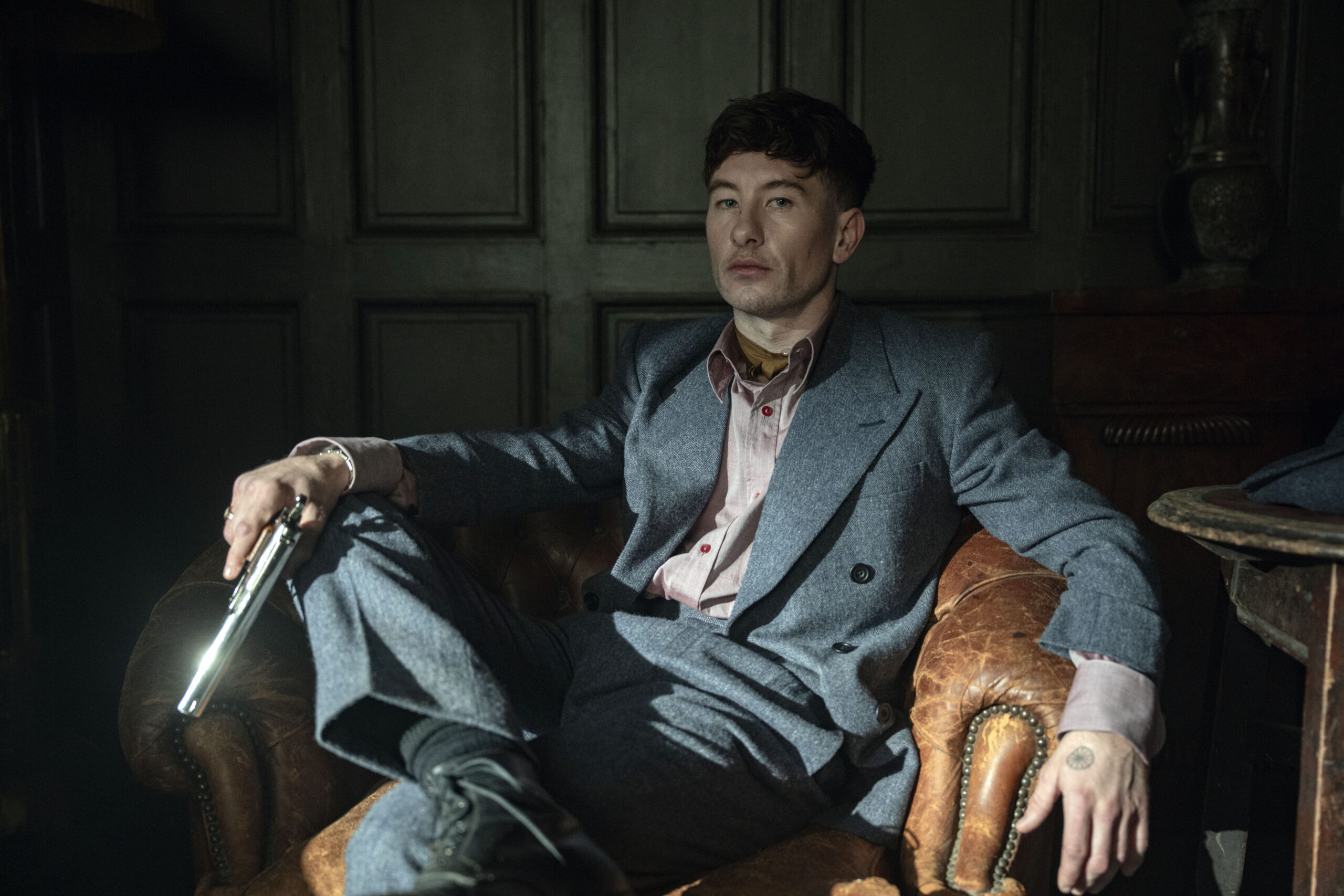 "Man in a gray suit sitting in a vintage leather armchair, holding a gun, in a dimly lit room with dark paneled walls, creating a tense and dramatic atmosphere."