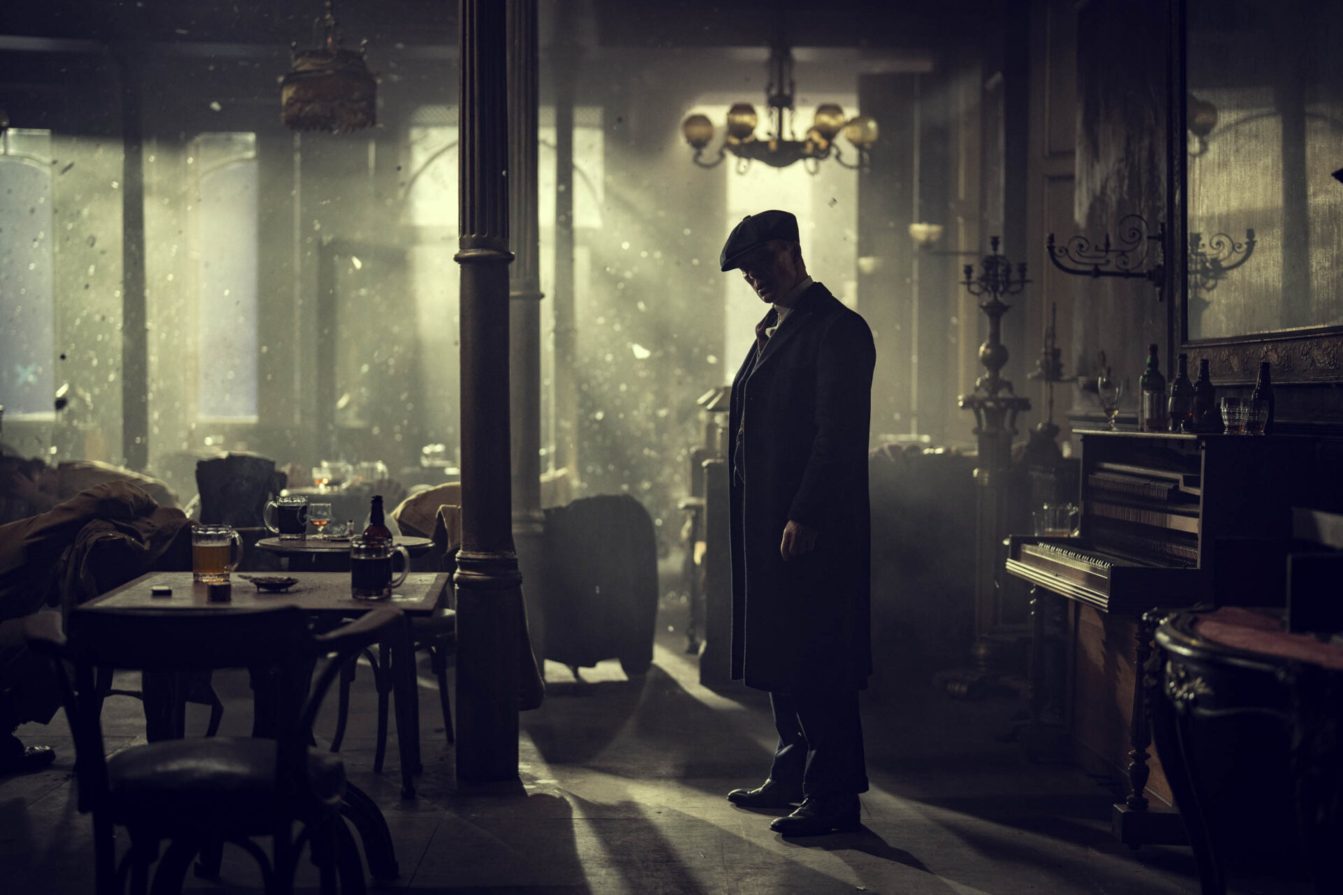 "A man in a long coat stands in a dimly lit, smoky vintage bar or pub, surrounded by empty tables, chairs, and an upright piano, with dramatic light streaming through the windows."