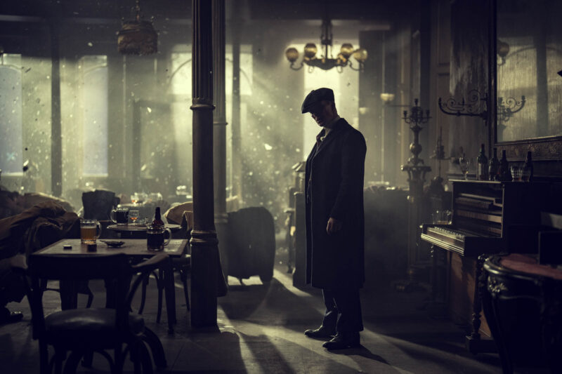 "A man in a long coat stands in a dimly lit, smoky vintage bar or pub, surrounded by empty tables, chairs, and an upright piano, with dramatic light streaming through the windows."